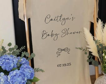 May include: A cream-colored baby shower welcome sign with the text "Welcome to Caitlyn's Baby Shower" and the date 07.19.25. The sign is decorated with a bow graphic and is held up by a wooden easel. Blue hydrangeas and pampas grass are in vases.
