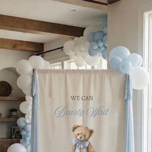 May include: A rectangular fabric sign featuring the text WE CAN Bearly Wait WELCOME TO July's and a central illustration of a brown teddy bear wearing a light blue ribbon. The sign is framed by light blue and white balloons on the top corners.