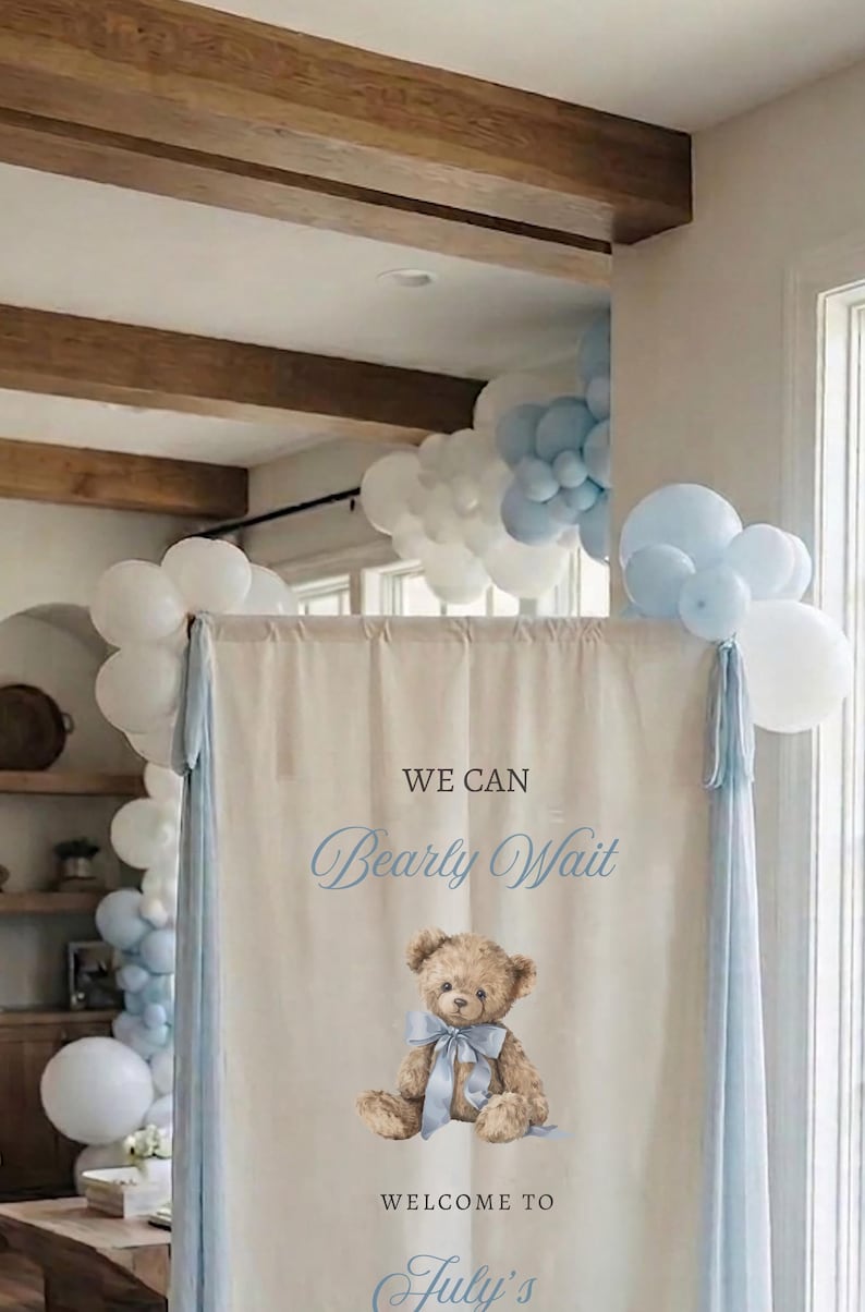 May include: A rectangular fabric sign featuring the text WE CAN Bearly Wait WELCOME TO July's and a central illustration of a brown teddy bear wearing a light blue ribbon. The sign is framed by light blue and white balloons on the top corners.
