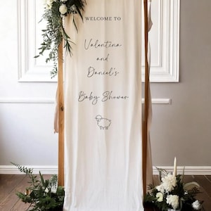 May include: A wooden frame with a cream-colored fabric banner. The banner reads "Welcome to Valentina and Daniel's Baby Shower" with a sheep illustration. The frame is decorated with greenery and white flowers.