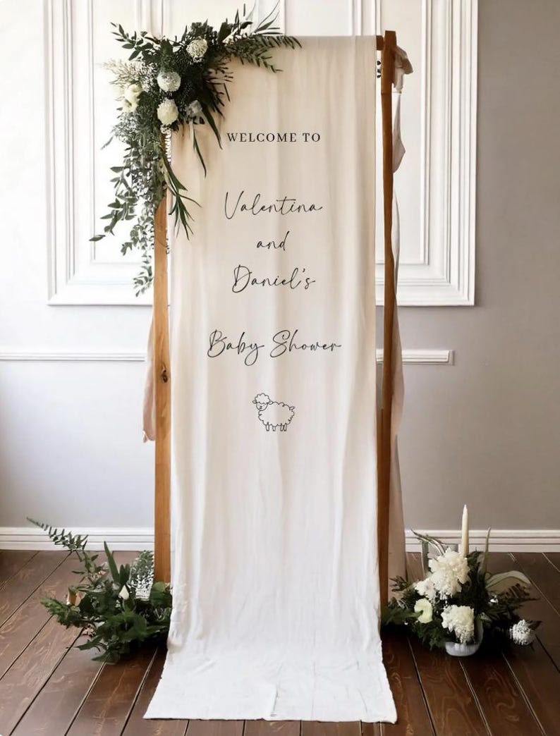 May include: A wooden frame with a cream-colored fabric banner. The banner reads "Welcome to Valentina and Daniel's Baby Shower" with a sheep illustration. The frame is decorated with greenery and white flowers.