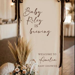 May include: A cream-colored baby shower sign with the text "Baby Riley is brewing" and "Welcome to Amelia Baby Shower" in brown script. The sign is decorated with brown ribbons, pampas grass, and a wicker baby carriage filled with flowers.