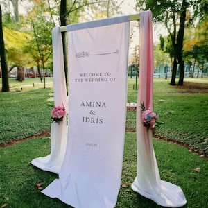 May include: White wedding ceremony archway decorated with pink and blue flowers. The sign reads "Welcome to the wedding of Amina & Idris 11.03.25"
