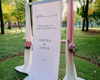 Pode incluir: Fundo branco para uma cerimônia de casamento com uma placa que diz "Bem-vindos ao casamento de Amina & Idris 11.03.25". O fundo é decorado com flores rosa e brancas.