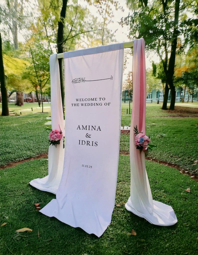 May include: White wedding ceremony archway decorated with pink and blue flowers. The sign reads "Welcome to the wedding of Amina & Idris 11.03.25"