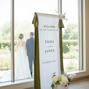 K&ouml;nnte beinhalten: Ein hohes, vertikales Hochzeits-Willkommensschild aus wei&szlig;em Stoff, das an einem Holzrahmen h&auml;ngt und den Text "WELCOME TO THE WEDDING OF EMMA and JAMES OCTOBER 8, 2025" tr&auml;gt. Es ist mit olivgr&uuml;nen Satinb&auml;ndern und wei&szlig;en Blumenarrangements verziert.