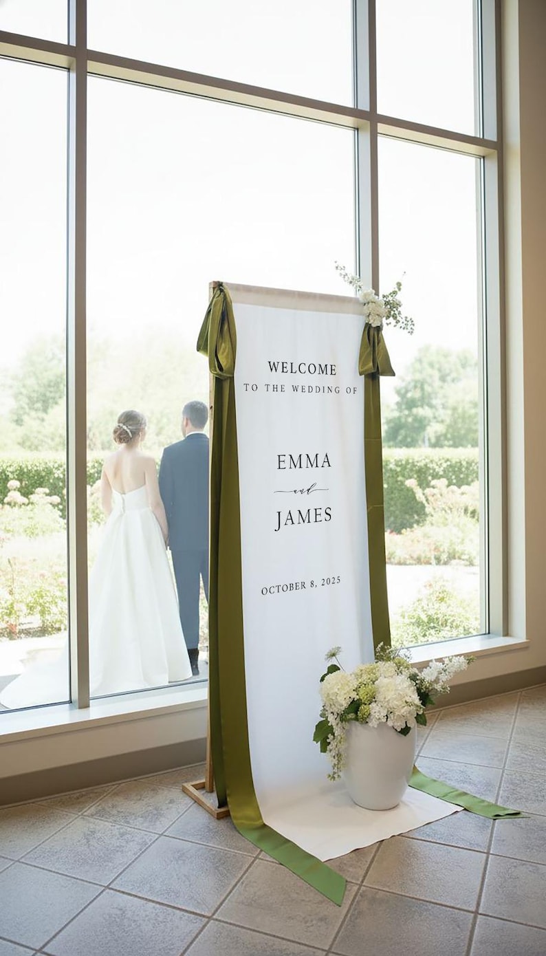 K&ouml;nnte beinhalten: Ein hohes, vertikales Hochzeits-Willkommensschild aus wei&szlig;em Stoff, das an einem Holzrahmen h&auml;ngt und den Text "WELCOME TO THE WEDDING OF EMMA and JAMES OCTOBER 8, 2025" tr&auml;gt. Es ist mit olivgr&uuml;nen Satinb&auml;ndern und wei&szlig;en Blumenarrangements verziert.