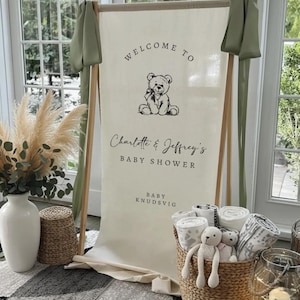 May include: A vertical cream-colored fabric banner suspended on a wooden frame, featuring a black line-art teddy bear illustration and the text: WELCOME TO Charlotte & Jeffrey's BABY SHOWER BABY KNUDSVIG. Sage green fabric drapes from the top corners.