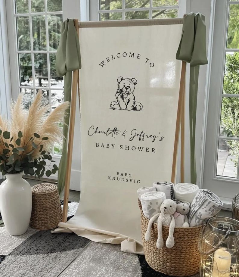 May include: A vertical cream-colored fabric banner suspended on a wooden frame, featuring a black line-art teddy bear illustration and the text: WELCOME TO Charlotte & Jeffrey's BABY SHOWER BABY KNUDSVIG. Sage green fabric drapes from the top corners.