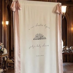 May include: Cream-colored banner with pink ribbon accents, featuring the text "a new chapter begins" above an open book illustration with flowers. Below, it reads "July's baby shower" and the date 12.05.2026.