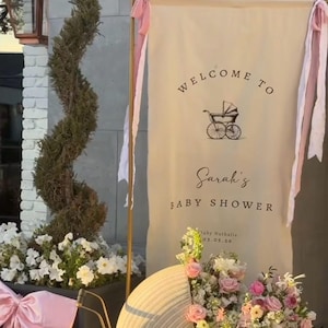 May include: A baby shower banner with the text "Welcome to Sarah's Baby Shower" and a vintage stroller illustration. The banner is decorated with pink ribbons and is next to a decorative stroller filled with flowers.