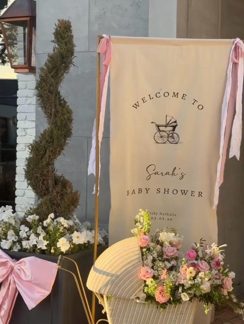 May include: A baby shower banner with the text "Welcome to Sarah's Baby Shower" and a vintage stroller illustration. The banner is decorated with pink ribbons and is next to a decorative stroller filled with flowers.