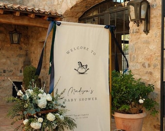 May include: A cream-colored banner with the words "WELCOME TO" and "Madison's BABY SHOWER" in black script. A rocking horse graphic is centered above the name. The banner is decorated with dark blue ribbons and a floral arrangement.