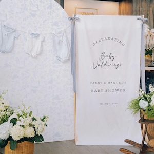 Op de afbeelding: Een wit rechthoekig stoffen spandoek met de tekst CELEBRATING Baby Valdiviezo FANNY & MANUEL'S BABY SHOWER January 11, 2026, opgehangen aan grijze linten. Daarnaast toont een ronde achtergrond lichtblauwe babykledingstukken.