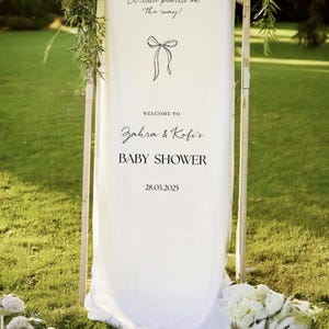 May include: A white fabric baby shower sign with the text "A little pearlis on the way!" and "Welcome to Zahra & Kofi's BABY SHOWER 28.03.2025". The sign is decorated with greenery and flowers, and is set on a green lawn.
