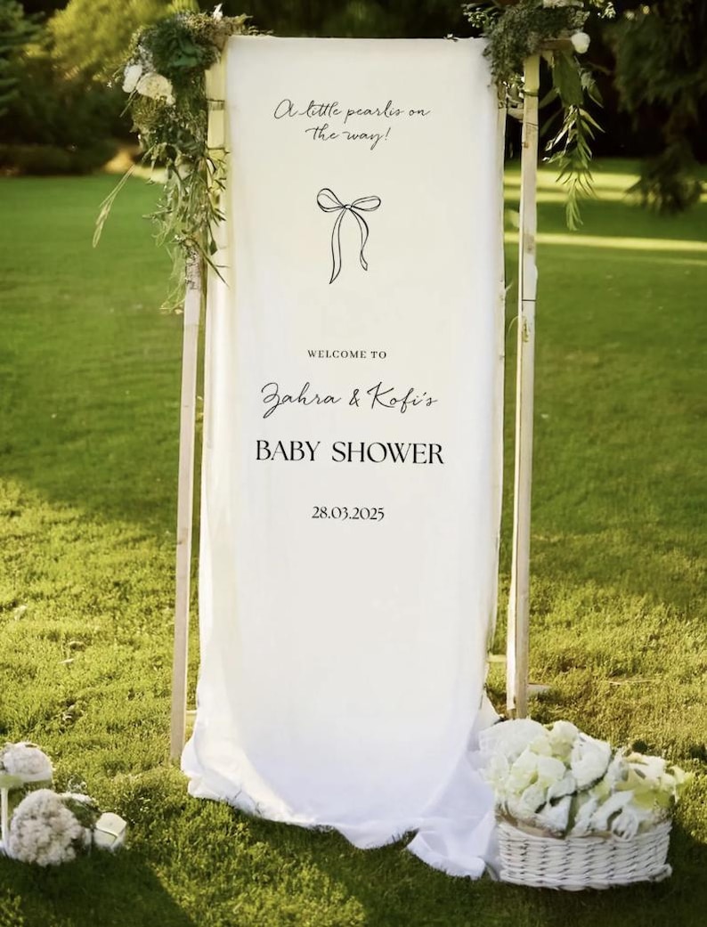 May include: A white fabric baby shower sign with the text "A little pearlis on the way!" and "Welcome to Zahra & Kofi's BABY SHOWER 28.03.2025". The sign is decorated with greenery and flowers, and is set on a green lawn.