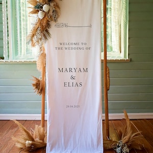 May include: White wedding welcome sign with calligraphy text that reads "Welcome to the wedding of Maryam & Elias 29.04.2025". The sign is hanging on a wooden frame with dried pampas grass and white flowers on either side.