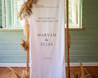 Pode incluir: Placa de boas-vindas de casamento branca com o texto "Bem-vindos ao casamento de Maryam & Elias 29.04.2025". A placa é decorada com capim-dos-pampas seco.