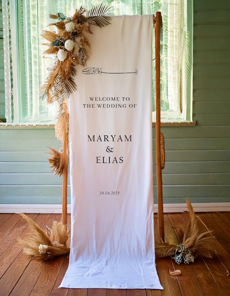 May include: White wedding welcome sign with calligraphy text that reads "Welcome to the wedding of Maryam & Elias 29.04.2025". The sign is hanging on a wooden frame with dried pampas grass and white flowers on either side.