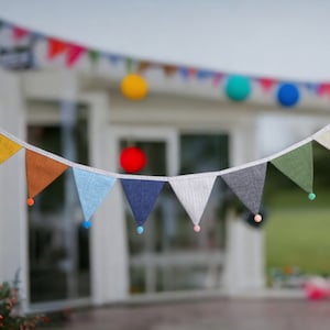 May include: A string of colourful fabric triangle flags with pom-poms hanging from a white string. The flags are in various shades of blue, green, yellow, and orange.