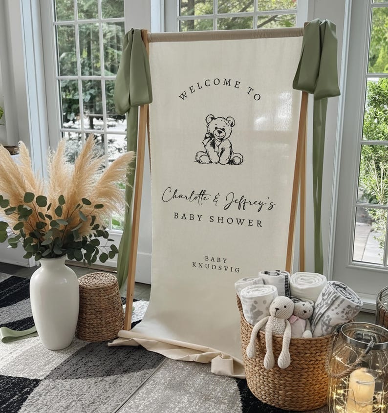 Może przedstawiać: Znak powitalny na baby shower z napisem "Welcome to Charlotte & Jeffrey's Baby Shower" i ilustracją misia. Wiklinowy kosz zawiera zwinięte koce i pluszaki. Widoczna jest r&oacute;wnież trawa pampas i biały wazon.