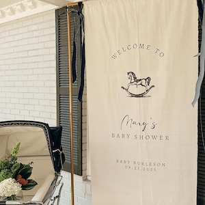 Może przedstawiać: Kremowy baner z napisem &bdquo;WELCOME TO Mary's BABY SHOWER&rdquo; i ilustracją konia na biegunach. Baner zawiera r&oacute;wnież tekst &bdquo;BABY BURLESON 09.21.2025&rdquo;. Zabytkowy w&oacute;zek dziecięcy znajduje się na pierwszym planie.