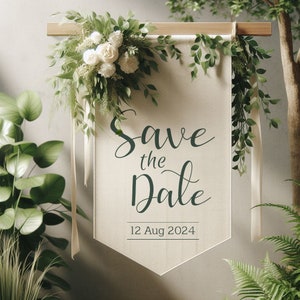 May include: A white banner with green foliage hanging from a wooden dowel. The banner reads "Save the Date" in cursive script with the date "12 Aug 2024" printed below.