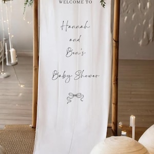 May include: A white fabric banner with the text "WELCOME TO Hannah and Ben's Baby Shower" in elegant script. The banner is framed by wooden poles and adorned with greenery. A decorative bow graphic is at the bottom.