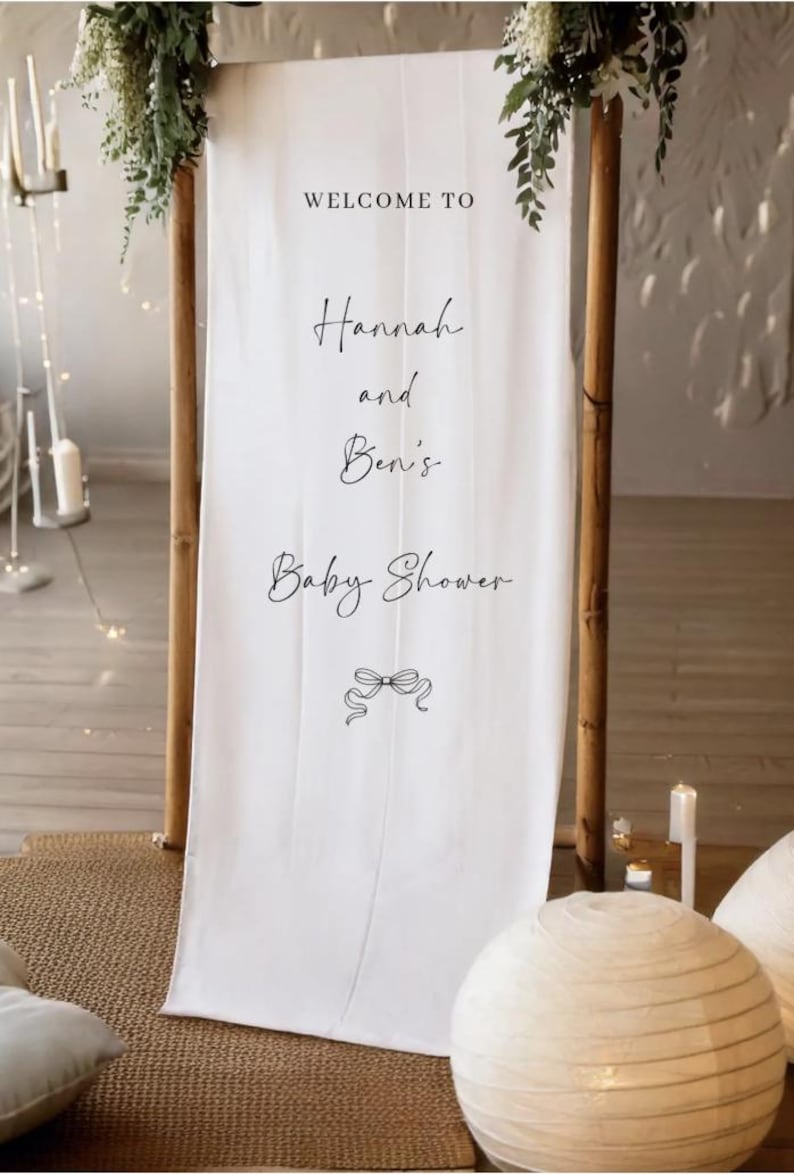 May include: A white fabric banner with the text "WELCOME TO Hannah and Ben's Baby Shower" in elegant script. The banner is framed by wooden poles and adorned with greenery. A decorative bow graphic is at the bottom.