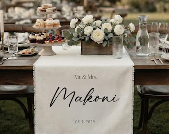 May include: A wedding table setting with a white linen runner bearing the names "Mr. & Mrs. Makoni" and the date 08.31.2025. The table is set with glassware, cutlery, and a floral arrangement in a wooden box. Desserts are displayed on a tiered stand.