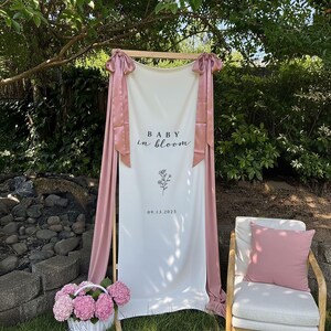 May include: A white banner with the text "BABY in bloom" and a floral illustration, framed by a wooden structure and draped with pink fabric. A white wicker basket with pink flowers sits in front.