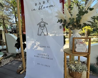 以下が含まれることがあります： 「BABY in bloom」と「WELCOME TO ALEENA'S BABY SPRINKLE 09-13-2025」と書かれた白いバナーのある木枠。バナーにはベビーオーバーオールの絵が描かれています。パンパスグラスと花の入った花瓶が木製のテーブルの上にあります。