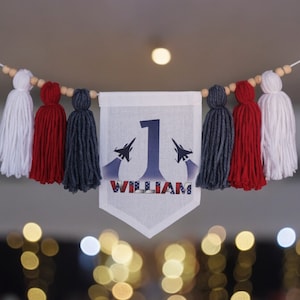 Könnte beinhalten: Ein weißes Banner mit roten, weißen und blauen Quasten hängt an einer Schnur. Das Banner hat ein blaues und weißes Flugzeugdesign mit der Nummer "1" und dem Namen "William" darauf gedruckt.
