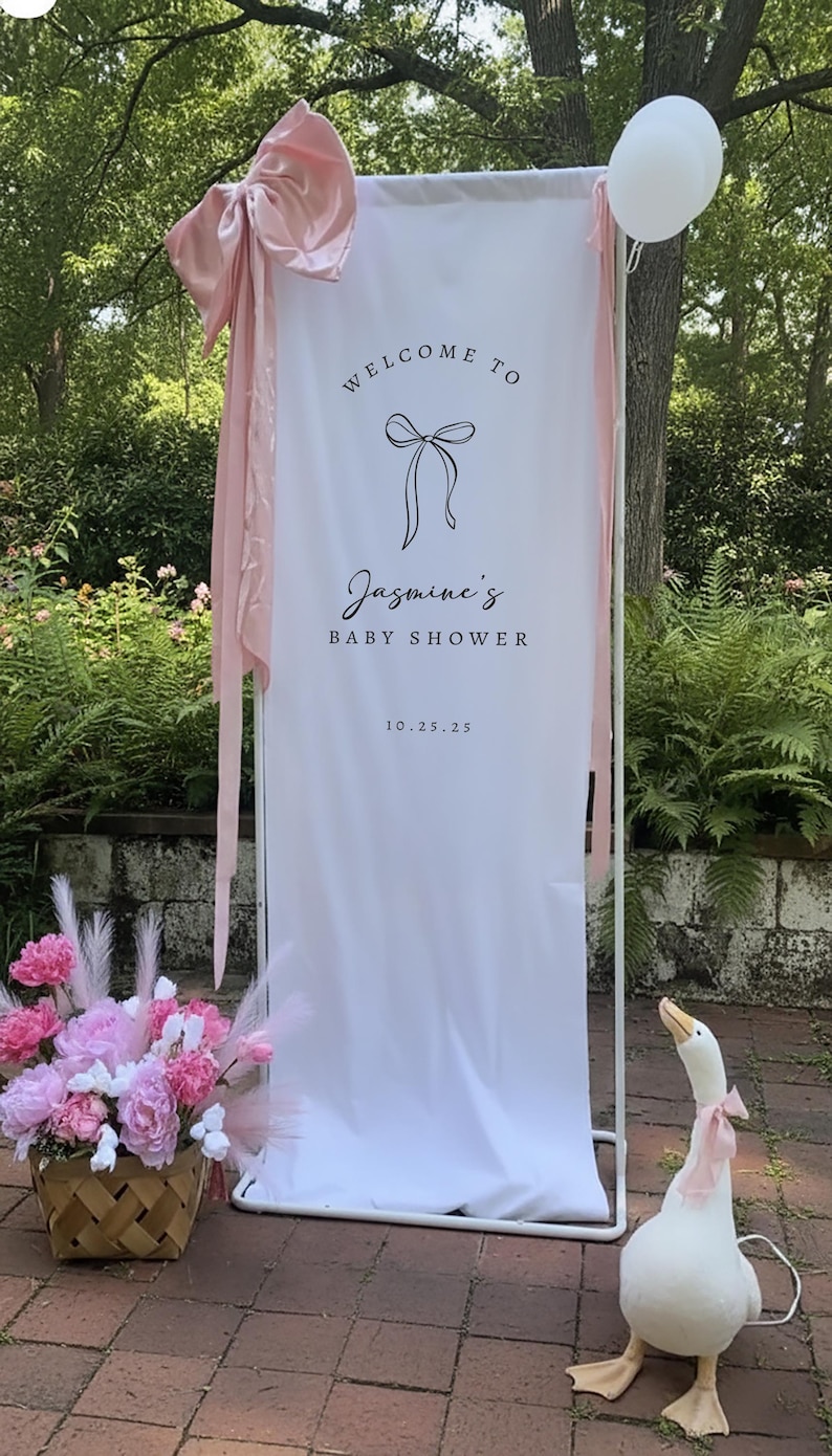 Może przedstawiać: Biały baner z napisem "Welcome to Jasmine's Baby Shower" i datą 10.25.25, ozdobiony r&oacute;żowymi kokardami i wstążkami. Obecny jest r&oacute;wnież kosz r&oacute;żowych i białych kwiat&oacute;w oraz biały balon. W pobliżu stoi dekoracyjna gęś z r&oacute;żową kokardą.