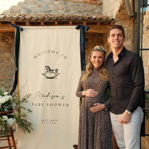 Może przedstawiać: Kremowy baner z granatowymi wstążkami, ogłaszający "Witamy na Baby Shower Madison" z ilustracją konia na biegunach. Para pozuje przed banerem. Widoczna jest r&oacute;wnież data 01-31-2026.
