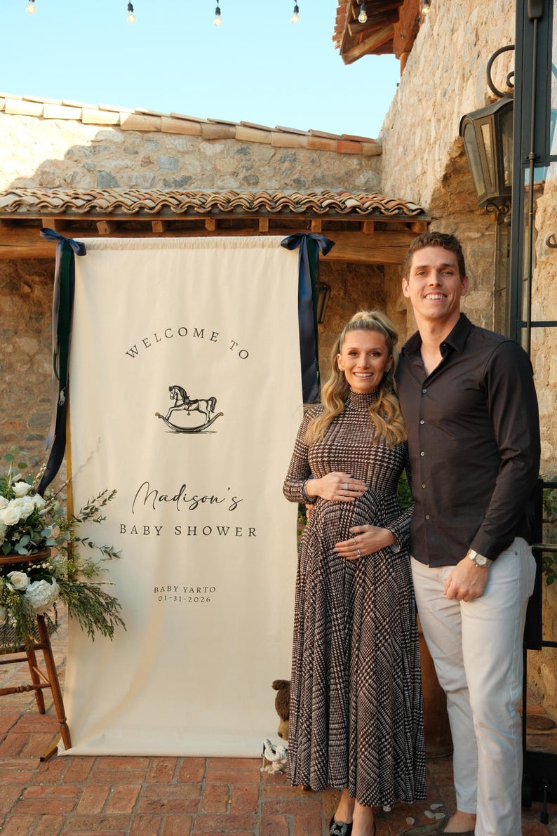 Może przedstawiać: Kremowy baner z granatowymi wstążkami, ogłaszający "Witamy na Baby Shower Madison" z ilustracją konia na biegunach. Para pozuje przed banerem. Widoczna jest r&oacute;wnież data 01-31-2026.