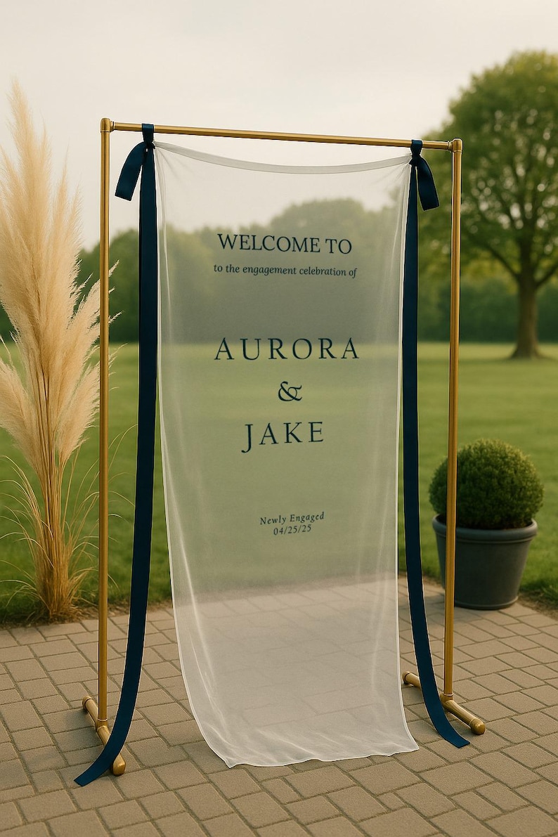 May include: A gold metal frame holds a sheer white fabric banner with navy blue ribbons. The banner reads "WELCOME TO the engagement celebration of AURORA & JAKE Newly Engaged 04/25/25". The scene is outdoors with greenery.