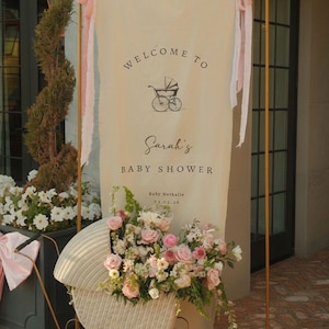 May include: A baby shower sign with the words "Welcome to Sarah's Baby Shower" and a vintage stroller illustration. The sign is decorated with pink ribbons and stands in front of a vintage stroller filled with flowers.