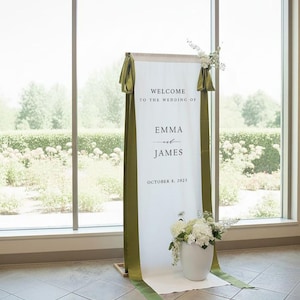 K&ouml;nnte beinhalten: Ein hohes, vertikales Hochzeits-Willkommensschild aus wei&szlig;em Stoff mit dem Text "WELCOME TO THE WEDDING OF EMMA and JAMES OCTOBER 8, 2025". Das Schild ist mit langen, olivgr&uuml;nen Satinb&auml;ndern an den Seiten und wei&szlig;en Blumenarrangements verziert.