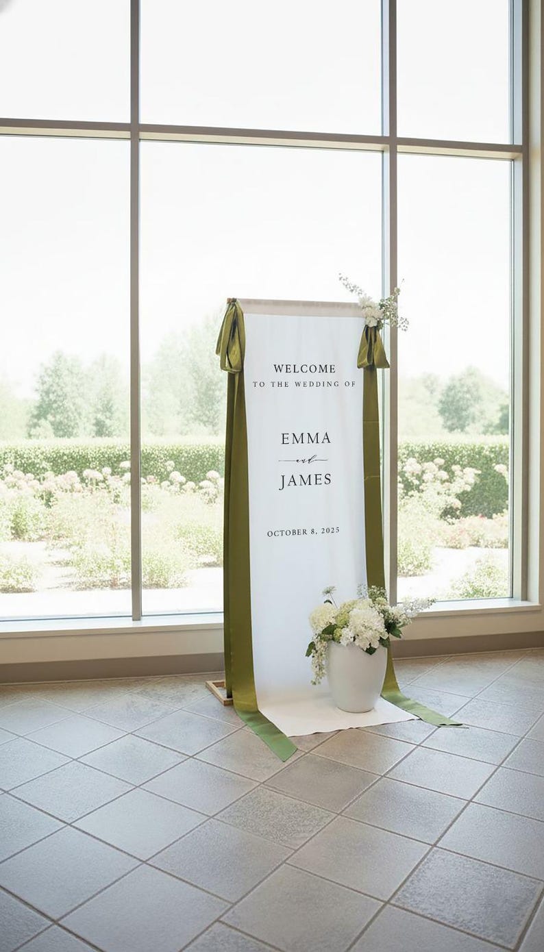 K&ouml;nnte beinhalten: Ein hohes, vertikales Hochzeits-Willkommensschild aus wei&szlig;em Stoff mit dem Text "WELCOME TO THE WEDDING OF EMMA and JAMES OCTOBER 8, 2025". Das Schild ist mit langen, olivgr&uuml;nen Satinb&auml;ndern an den Seiten und wei&szlig;en Blumenarrangements verziert.