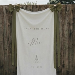 May include: A white fabric banner with the text "HAPPY BIRTHDAY Mia" in black. A cake illustration and the date 05.11.2025 are below. Olive green bows and ribbons are attached to the top corners, hanging against a wooden fence.