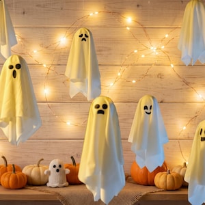 Pode incluir: Uma coleção de decorações de fantasmas de tecido branco com traços faciais pretos, alguns suspensos e outros sobre abóboras laranja. Um fio de luzes brancas quentes está colocado atrás dos fantasmas, iluminando a cena.