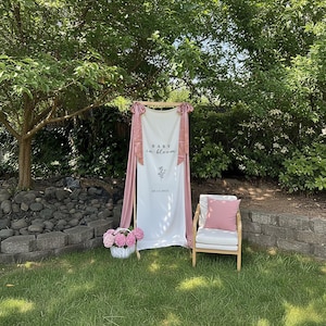 May include: A white banner with the text "BABY in bloom" and a floral design, draped with pink fabric. A wooden chair with a pink pillow and a basket of pink flowers are in front of a green background.
