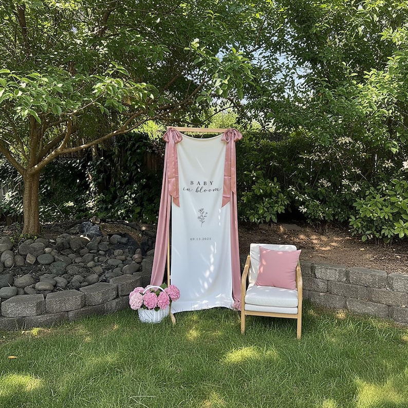May include: A white banner with the text "BABY in bloom" and a floral design, draped with pink fabric. A wooden chair with a pink pillow and a basket of pink flowers are in front of a green background.