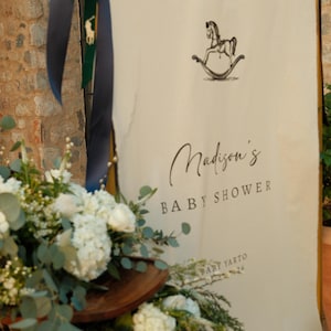 Może przedstawiać: Kremowy baner z napisem "WELCOME TO" i "Madison's BABY SHOWER" czarnym pismem. Ilustracja konia na biegunach znajduje się nad tekstem baby shower. Kompozycja kwiatowa z białymi kwiatami i zielenią znajduje się na pierwszym planie.