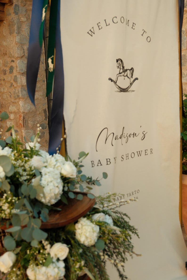 Może przedstawiać: Kremowy baner z napisem "WELCOME TO" i "Madison's BABY SHOWER" czarnym pismem. Ilustracja konia na biegunach znajduje się nad tekstem baby shower. Kompozycja kwiatowa z białymi kwiatami i zielenią znajduje się na pierwszym planie.