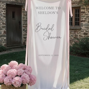 Op de afbeelding: Een witte stoffen banner met roze linten kondigt "Welcome to Sheldon's Bridal Shower" aan met de datum 12 september 2025. Een mand met roze bloemen staat voor de banner, tegen een stenen gebouw.