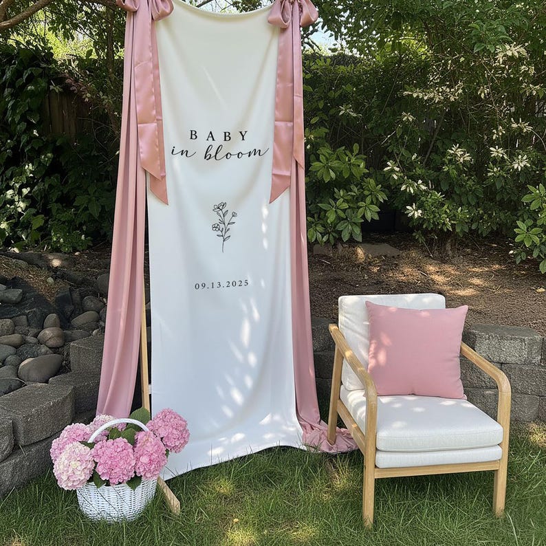 May include: A white banner with the text "BABY in BLOOM" and the date 09.13.2025, decorated with pink fabric and bows. A white wicker basket filled with pink hydrangeas sits on the grass, near a wooden chair with a white cushion and pink pillow.