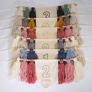 May include: A white fabric banner with the numbers 1, 2, and 3 embroidered on it. The banner is decorated with colorful yarn tassels and wooden beads. The bottom banner has the name "Emma" embroidered on it.