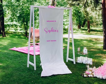 May include: White backdrop with a pink sign that says "Welcome to Sophia's Baby Shower" set up in a grassy outdoor area.